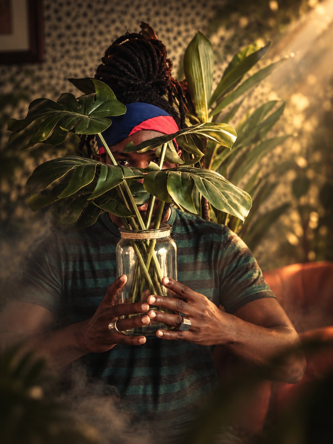 A man's face obscured behind a jar of monstera leaves, backlit by warm window light